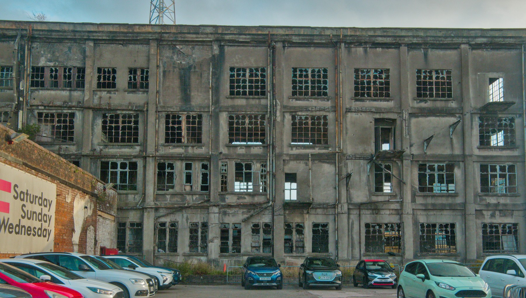 Weathered multi-storey industrial building at Sentinel Works with many broken-pane windows, seen above a row of parked cars.