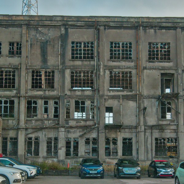 Weathered multi-storey industrial building at Sentinel Works with many broken-pane windows, seen above a row of parked cars.