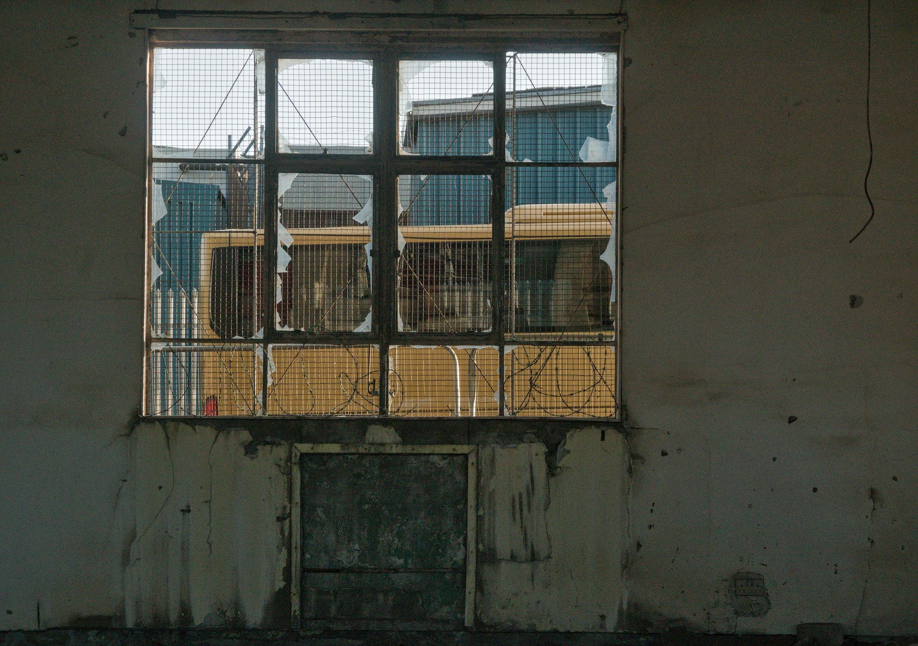 Broken, wire-mesh-covered window in a stained industrial wall, with yellow and blue structures visible beyond the shattered panes.