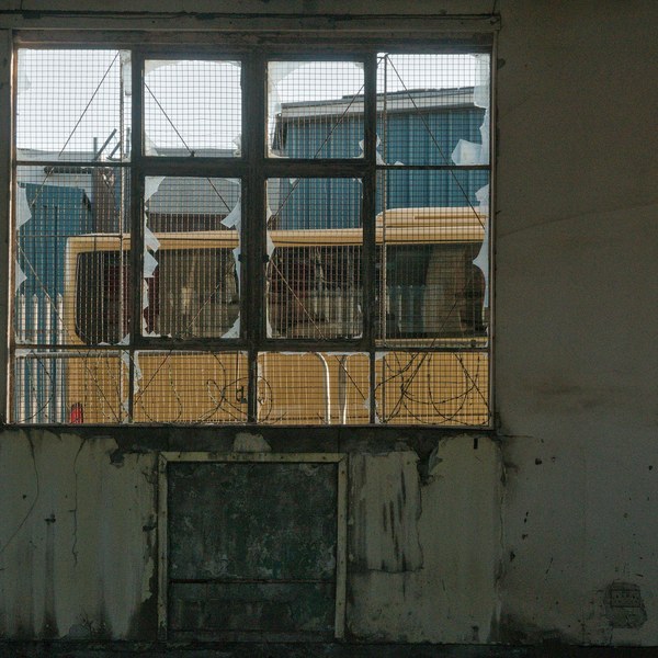Broken, wire-mesh-covered window in a stained industrial wall, with yellow and blue structures visible beyond the shattered panes.