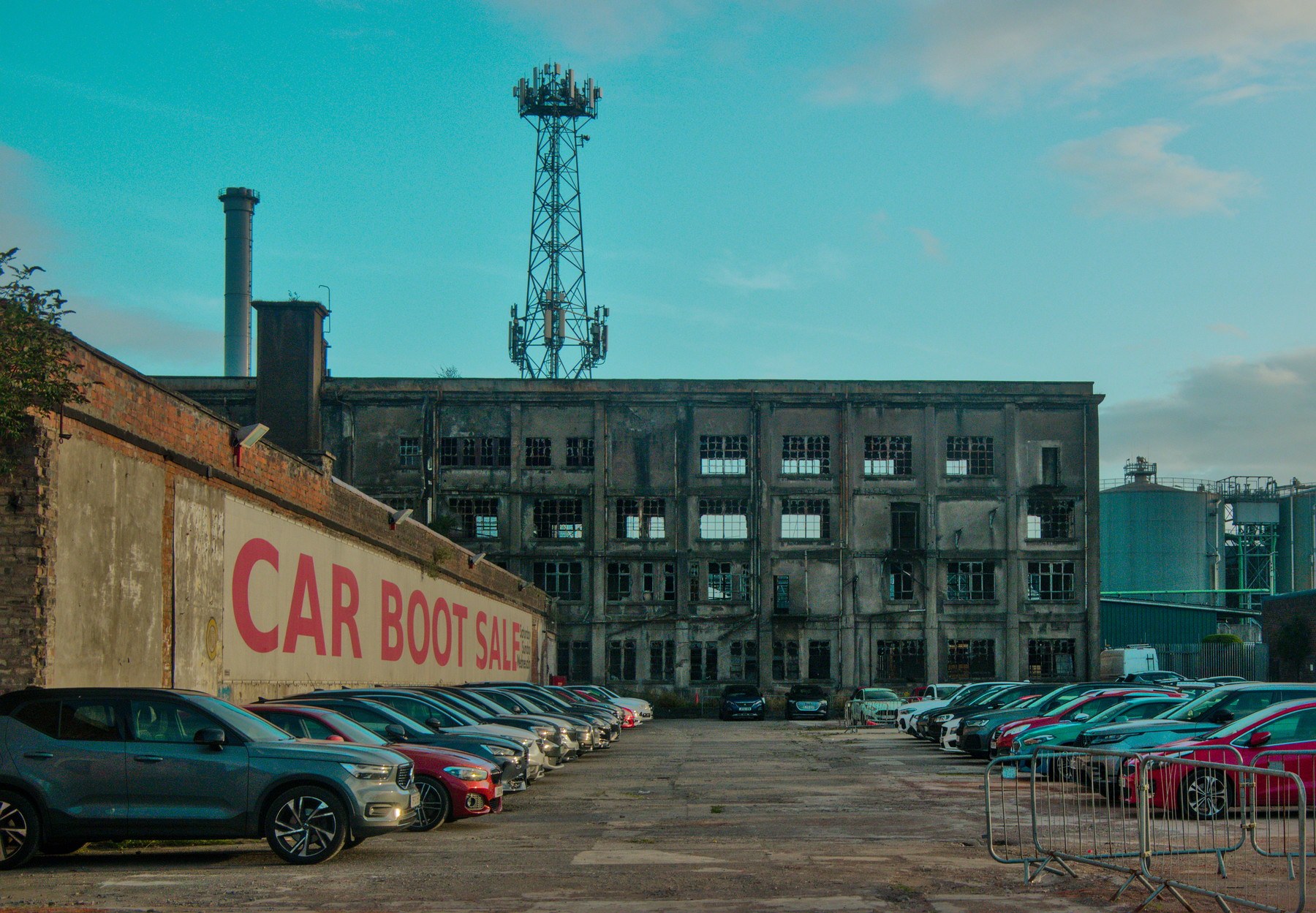 Rows of parked cars fill a car boot sale yard in front of the weathered Sentinel Works industrial building, with a tall communications mast rising above it.
