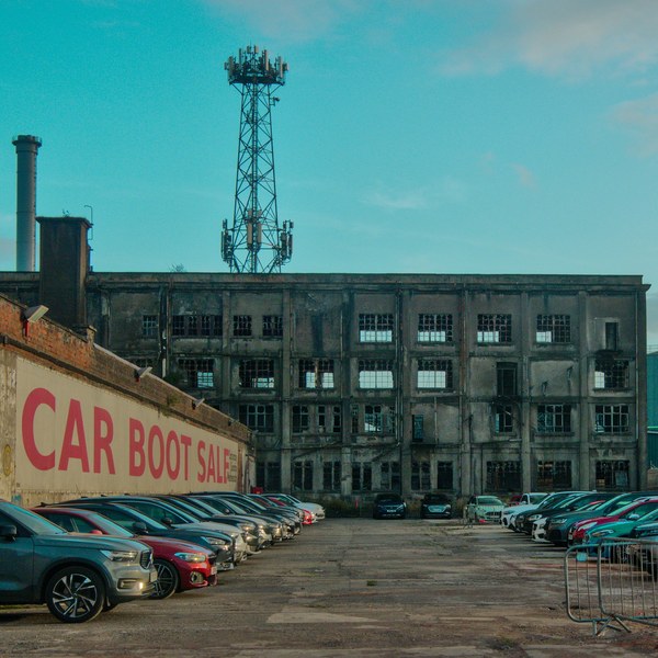 Rows of parked cars fill a car boot sale yard in front of the weathered Sentinel Works industrial building, with a tall communications mast rising above it.