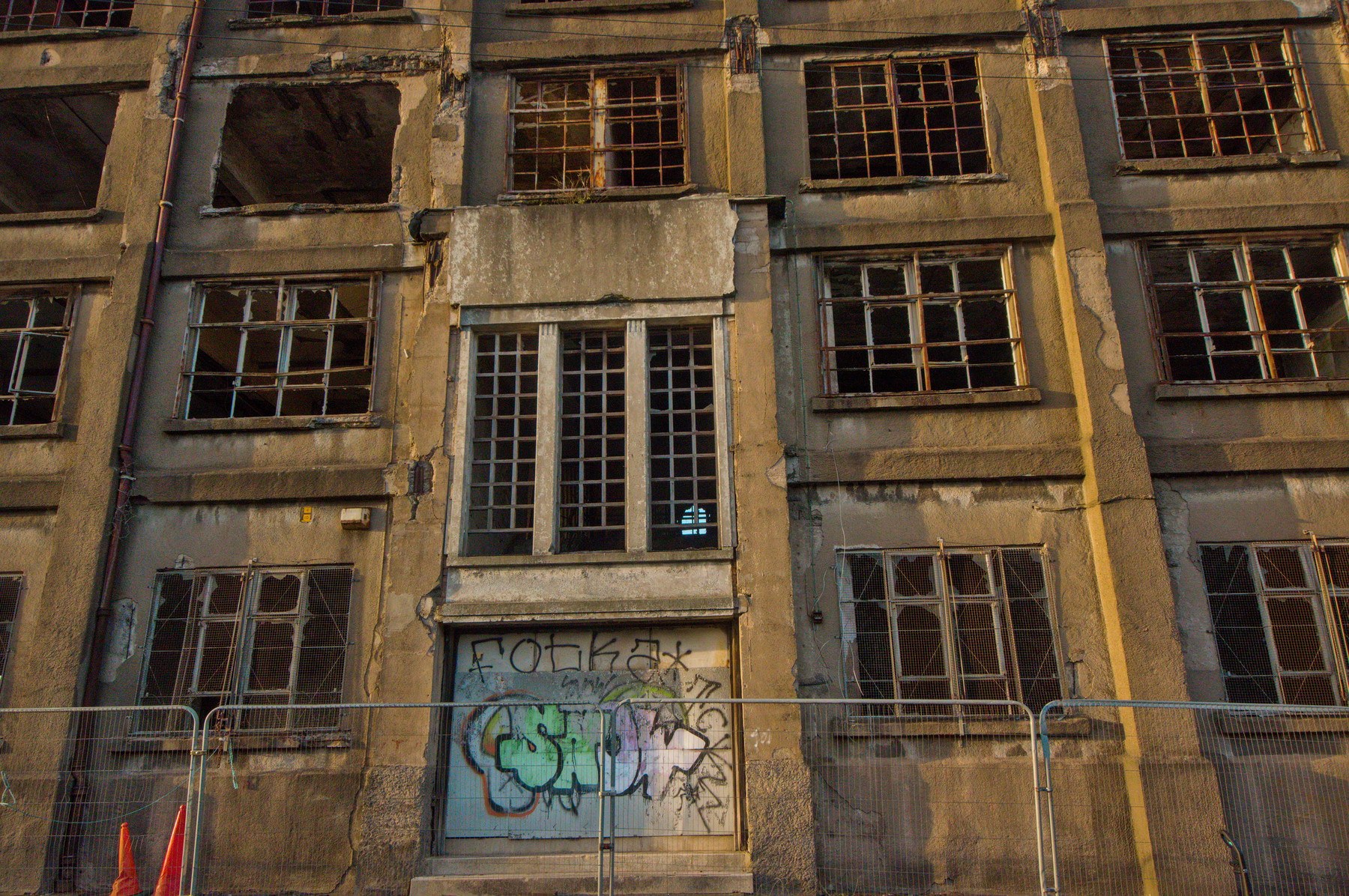 Weathered multi-storey facade of Sentinel Works in Glasgow, with broken-paned windows, rusted metal grilles, graffiti on a boarded ground-floor opening, and temporary barriers along the pavement.