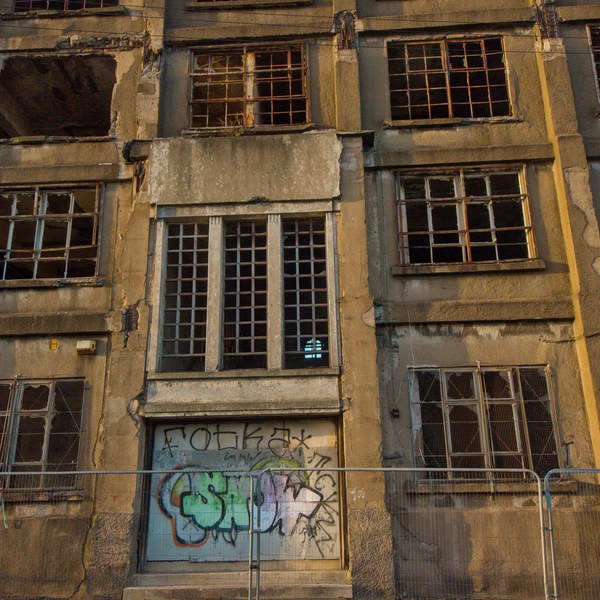 Weathered multi-storey facade of Sentinel Works in Glasgow, with broken-paned windows, rusted metal grilles, graffiti on a boarded ground-floor opening, and temporary barriers along the pavement.