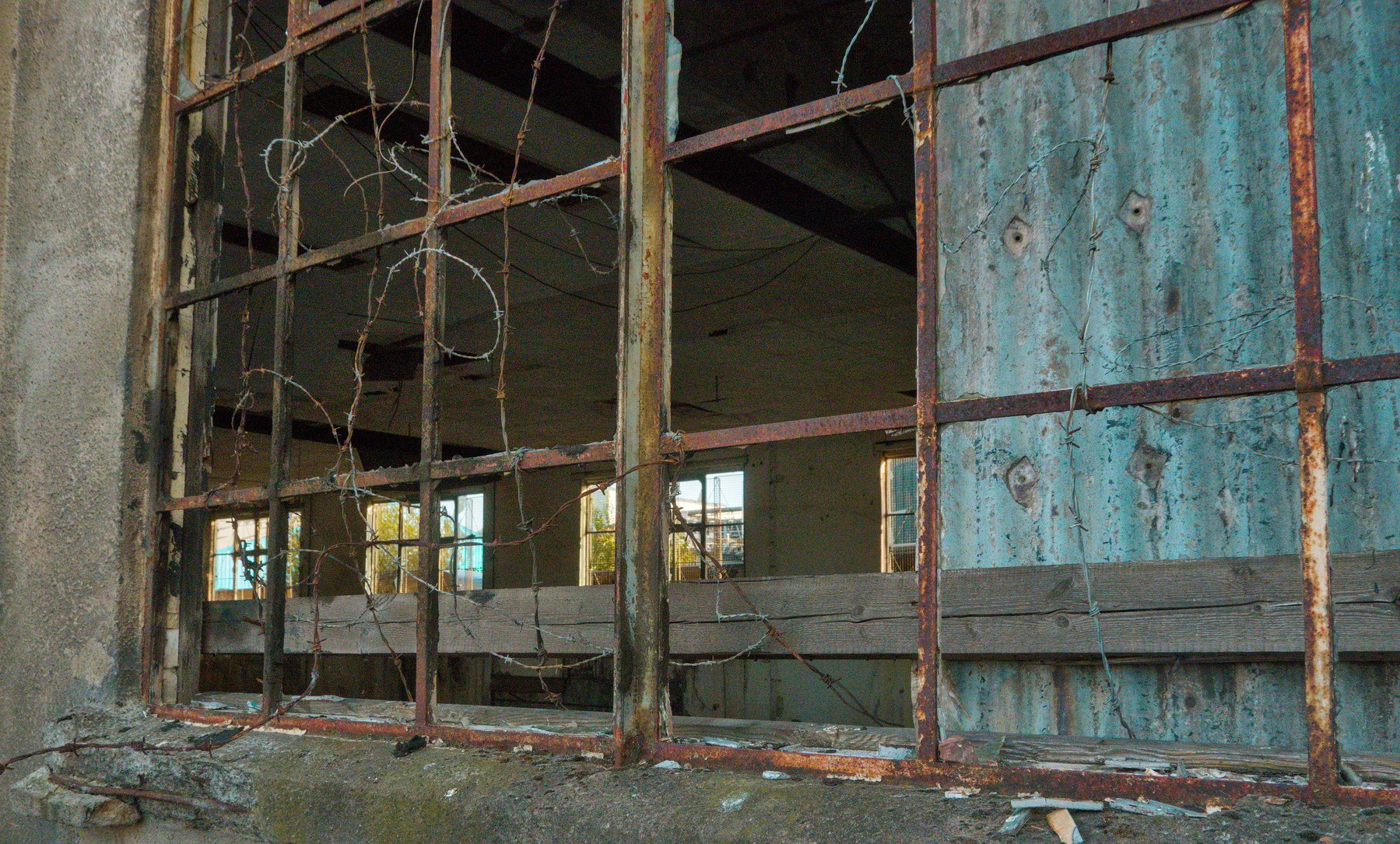 Broken, rusted window framework at Sentinel Works, revealing an empty industrial interior with peeling blue wall panels and hanging wires.