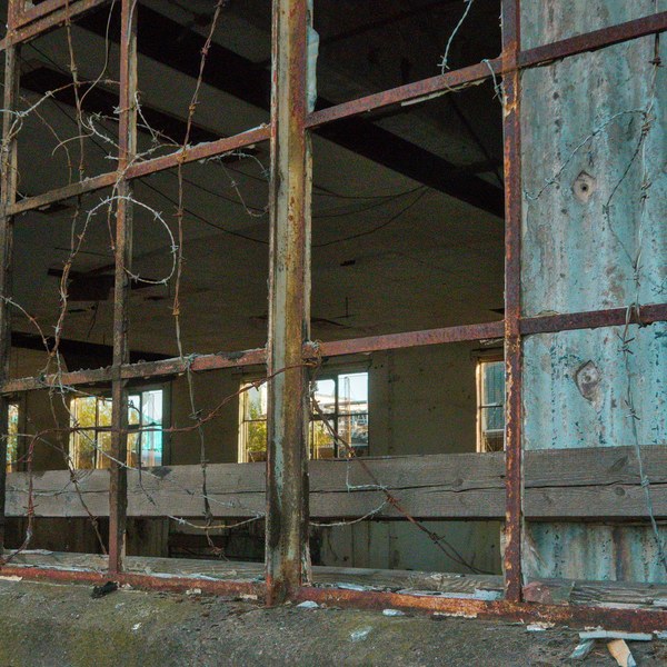 Broken, rusted window framework at Sentinel Works, revealing an empty industrial interior with peeling blue wall panels and hanging wires.