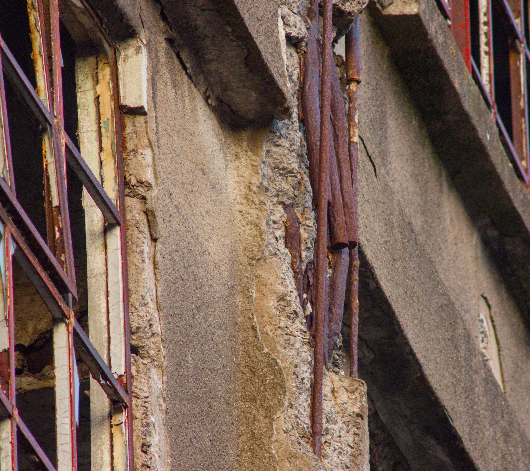 Close view of a deteriorating industrial building facade, with cracked concrete, exposed aggregate, rusted metal pipes and a red-painted steel frame at left.