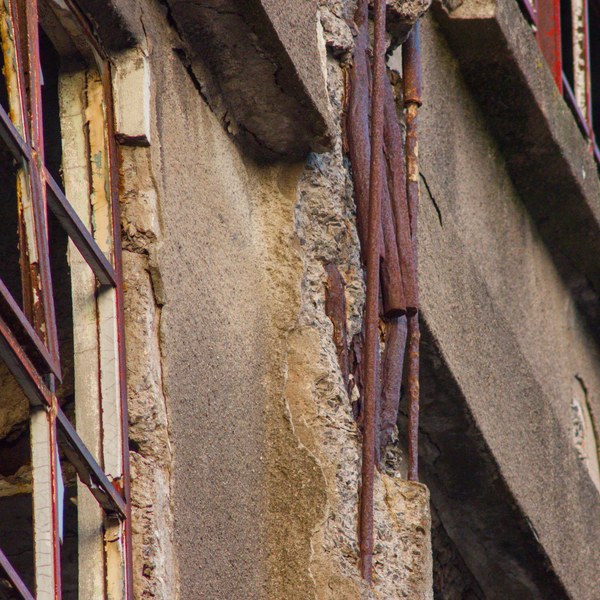 Close view of a deteriorating industrial building facade, with cracked concrete, exposed aggregate, rusted metal pipes and a red-painted steel frame at left.