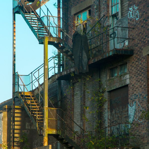Rusting external fire escape climbs a graffiti-marked brick building with boarded openings, turquoise-painted panes and weeds growing along the landings.