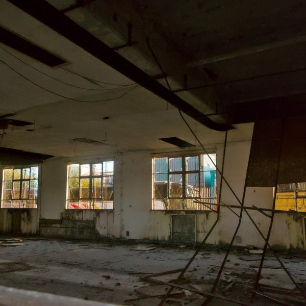 Dim, derelict interior at Sentinel Works, with debris on the concrete floor, hanging cables, broken ceiling panels and rows of industrial windows.