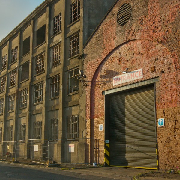 Weathered brick and concrete industrial building at Sentinel Works, with rows of broken-pane windows and a shuttered entrance beneath a faded sign.
