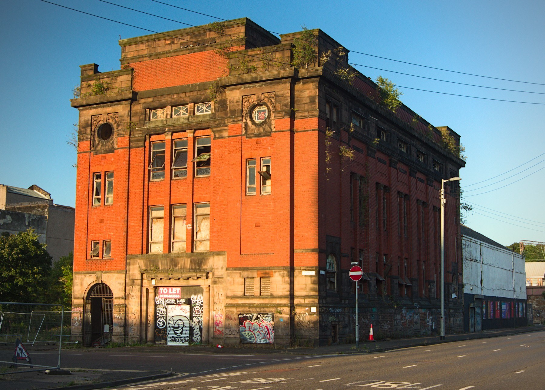 Sunlit red-brick former industrial building with boarded and damaged panes, circular wall openings, graffiti-covered ground-floor doors, a To Let sign and overhead power lines beside an empty road.
