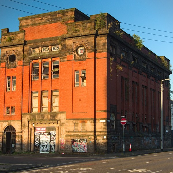 Sunlit red-brick former industrial building with boarded and damaged panes, circular wall openings, graffiti-covered ground-floor doors, a To Let sign and overhead power lines beside an empty road.