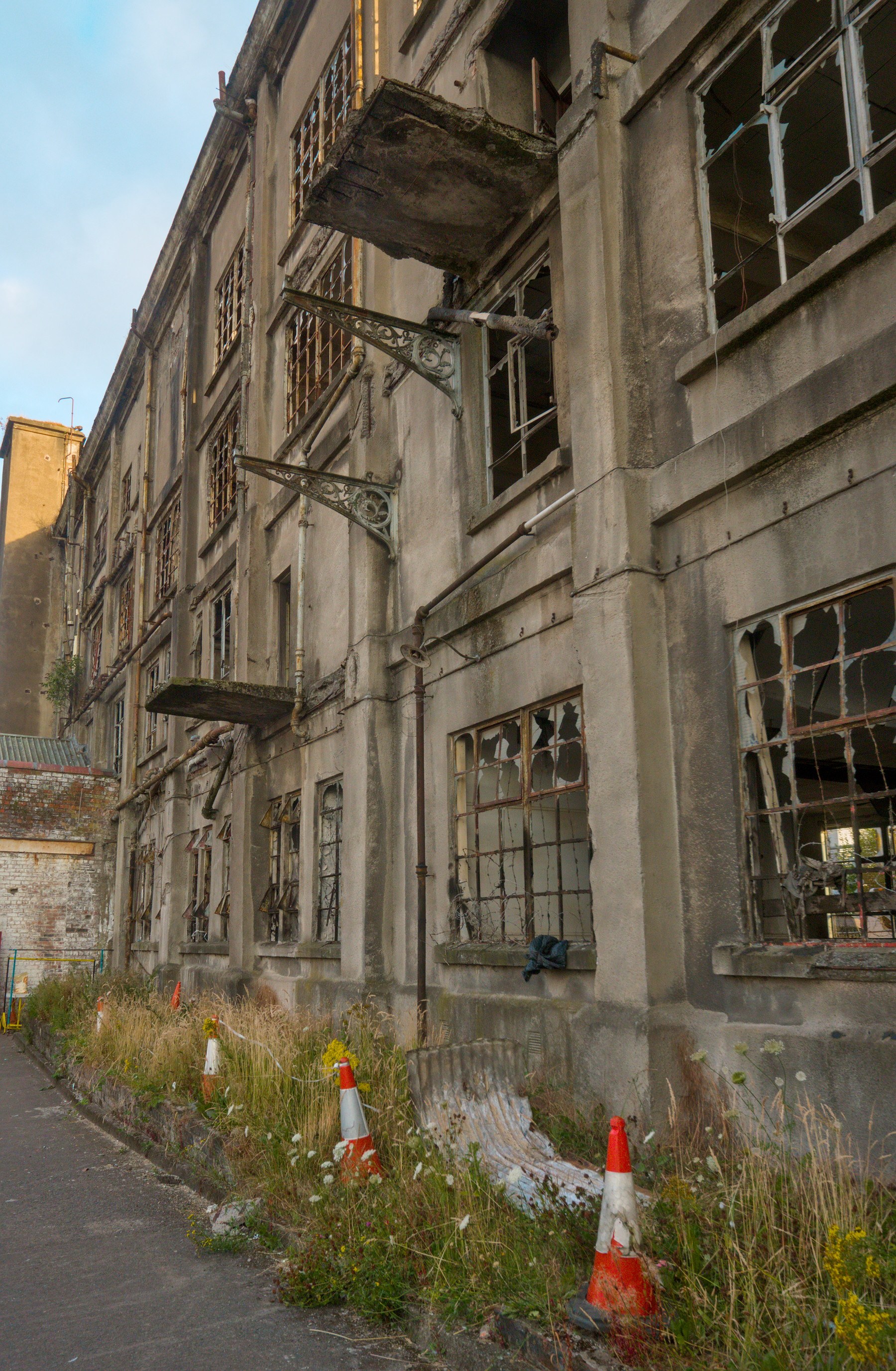 Weathered exterior of Sentinel Works with shattered multi-pane windows, rusted metal brackets, broken platforms, overgrown weeds and orange traffic cones.