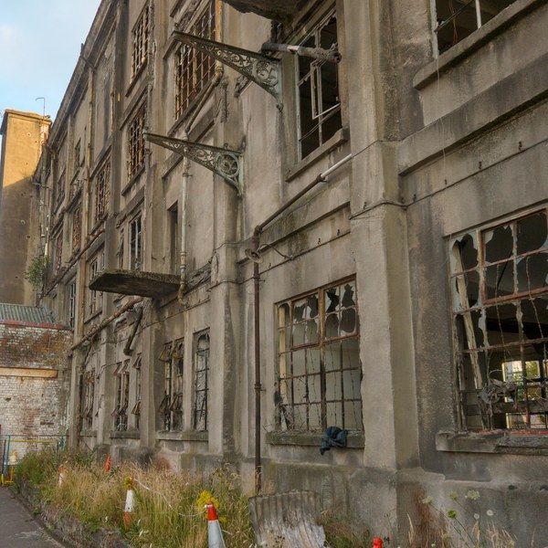 Weathered exterior of Sentinel Works with shattered multi-pane windows, rusted metal brackets, broken platforms, overgrown weeds and orange traffic cones.
