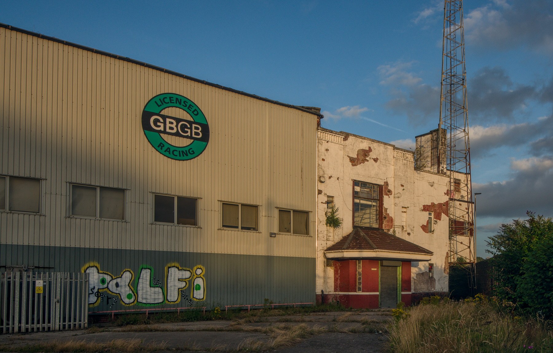 Derelict industrial buildings at Shawfield Stadium, including a corrugated facade with a GBGB Racing sign, graffiti, a boarded entrance and a tall metal tower beneath a blue evening sky.