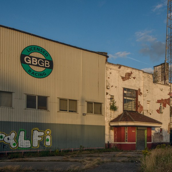 Derelict industrial buildings at Shawfield Stadium, including a corrugated facade with a GBGB Racing sign, graffiti, a boarded entrance and a tall metal tower beneath a blue evening sky.