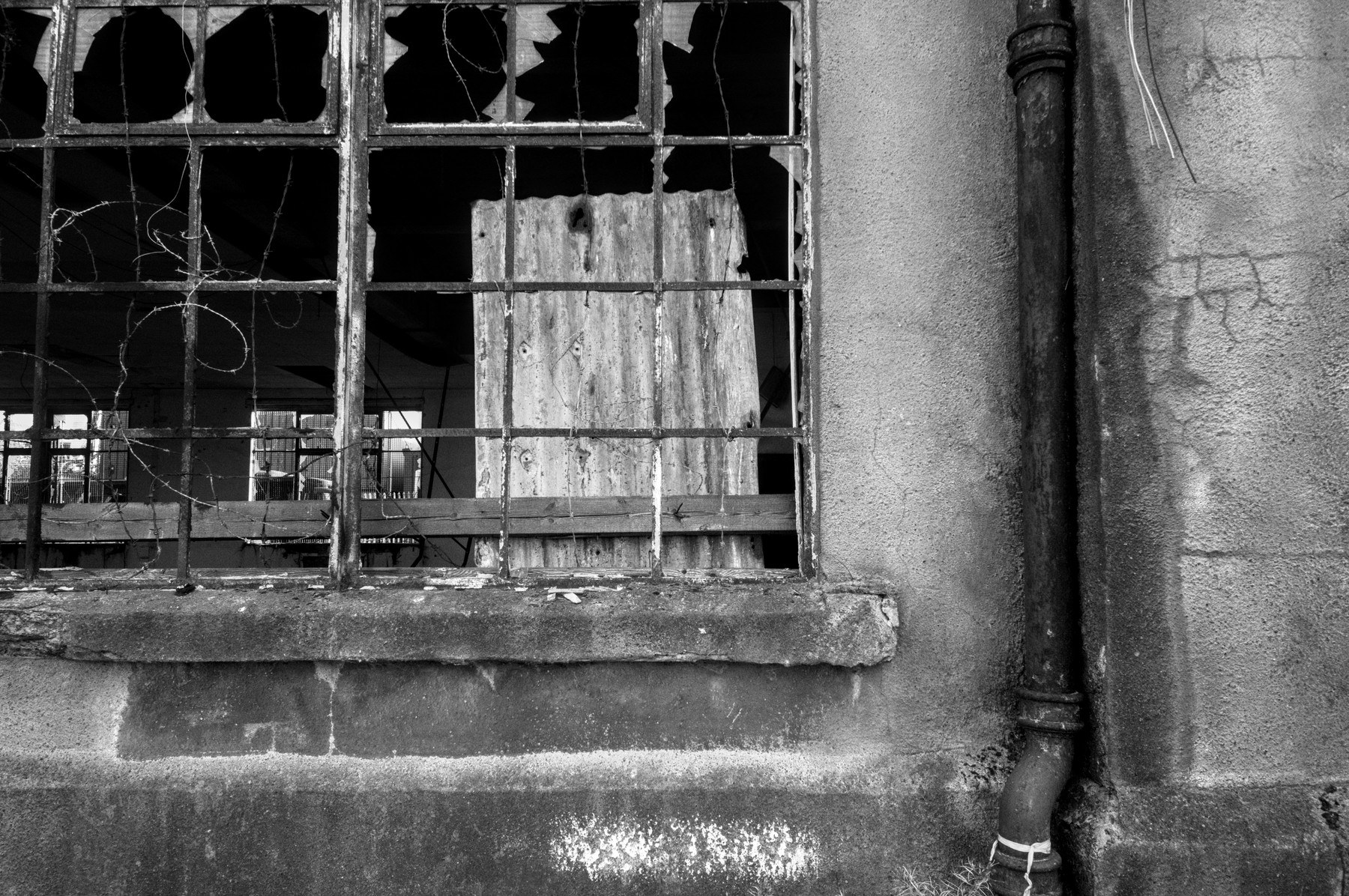 Broken-paned metal-framed window with a boarded opening and exposed wires beside a drainpipe on the weathered wall of Sentinel Works.