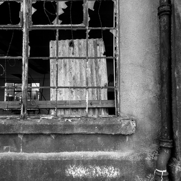 Broken-paned metal-framed window with a boarded opening and exposed wires beside a drainpipe on the weathered wall of Sentinel Works.