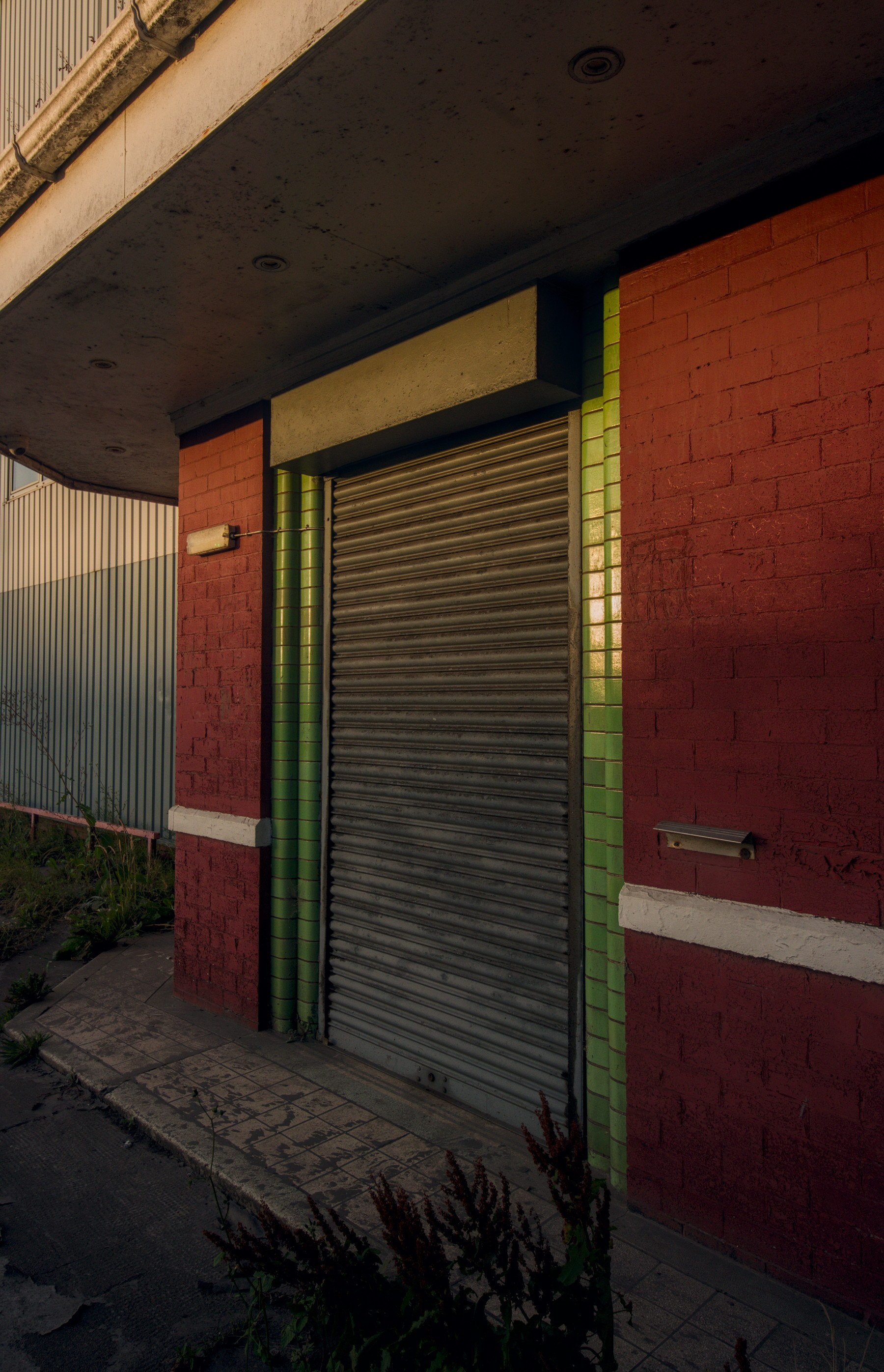 Closed metal roller shutter set in a red-brick industrial building, framed by glossy green tiles beneath a weathered concrete canopy.