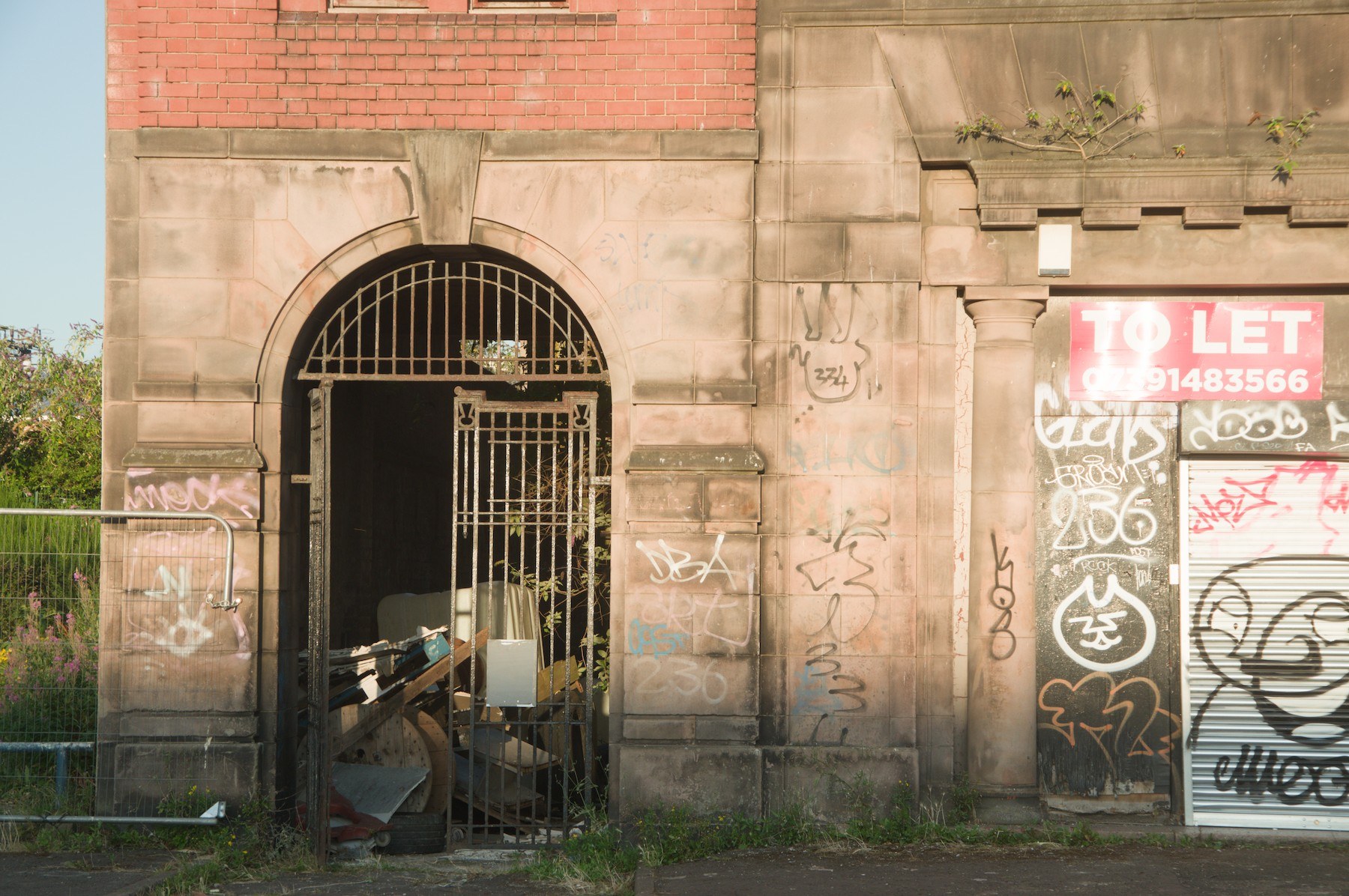 Graffiti-covered stone building exterior with a barred arched gateway, a pink To Let sign, and a shuttered opening at right.