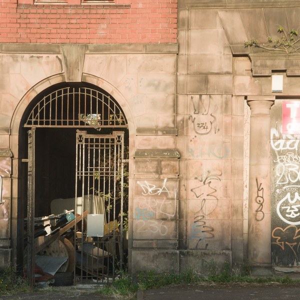 Graffiti-covered stone building exterior with a barred arched gateway, a pink To Let sign, and a shuttered opening at right.