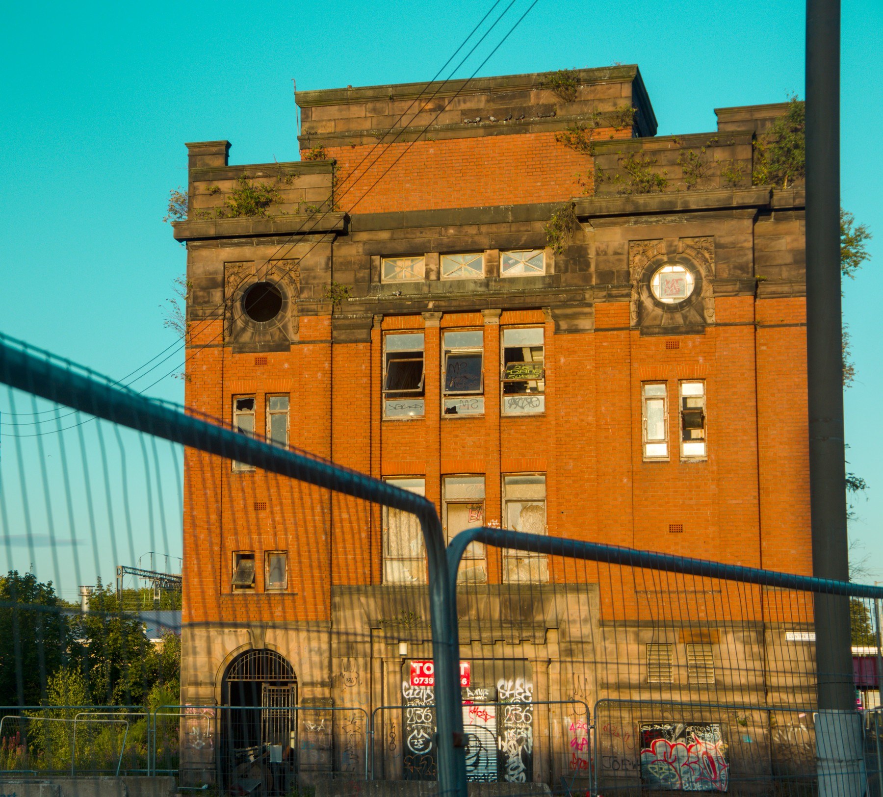 Orange-brick industrial building with stone details and circular openings, viewed behind a curved metal security fence; graffiti marks the ground-floor entrance beneath overhead wires.
