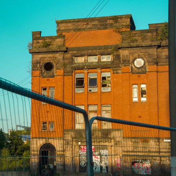 Orange-brick industrial building with stone details and circular openings, viewed behind a curved metal security fence; graffiti marks the ground-floor entrance beneath overhead wires.