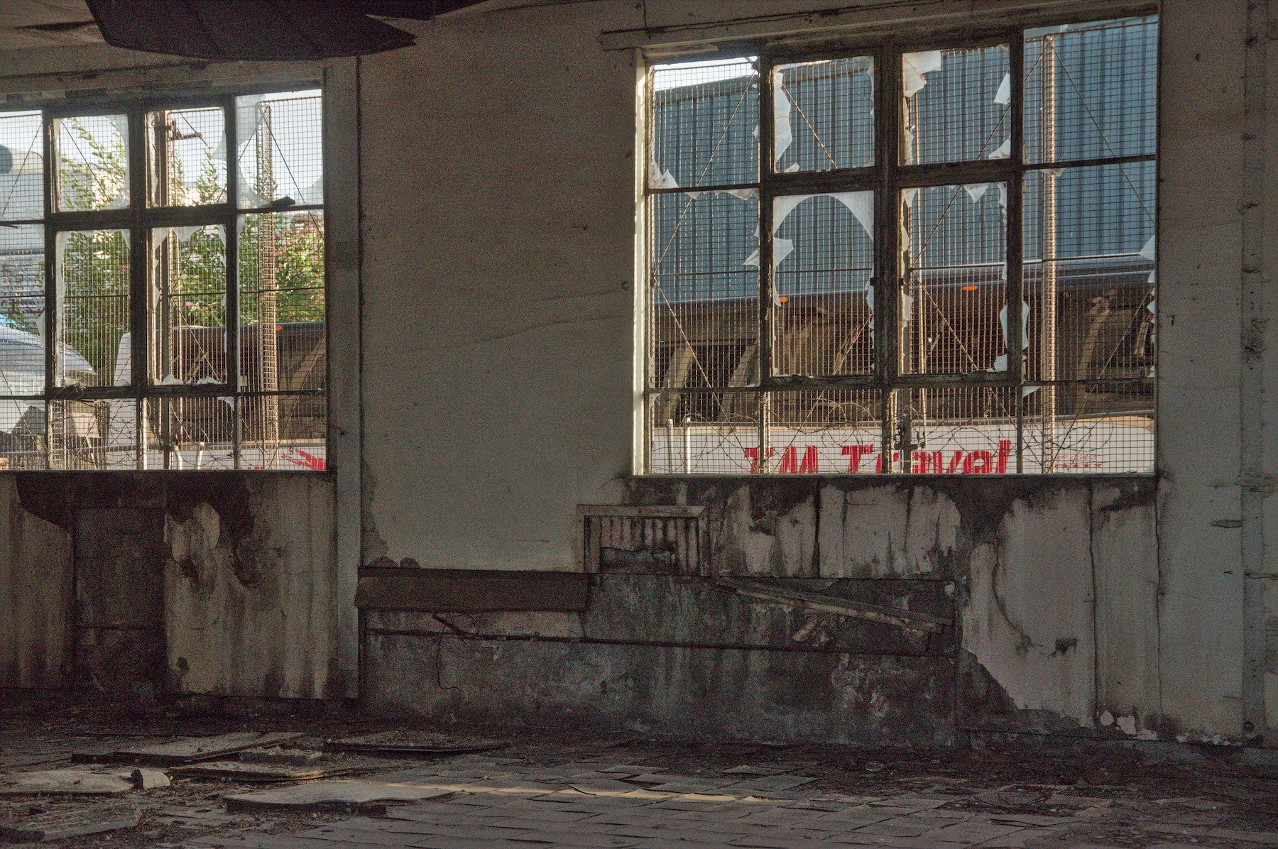 Derelict industrial interior with cracked plaster walls, debris-strewn floor and two large wire-mesh windows with shattered panes.