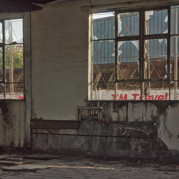 Derelict industrial interior with cracked plaster walls, debris-strewn floor and two large wire-mesh windows with shattered panes.