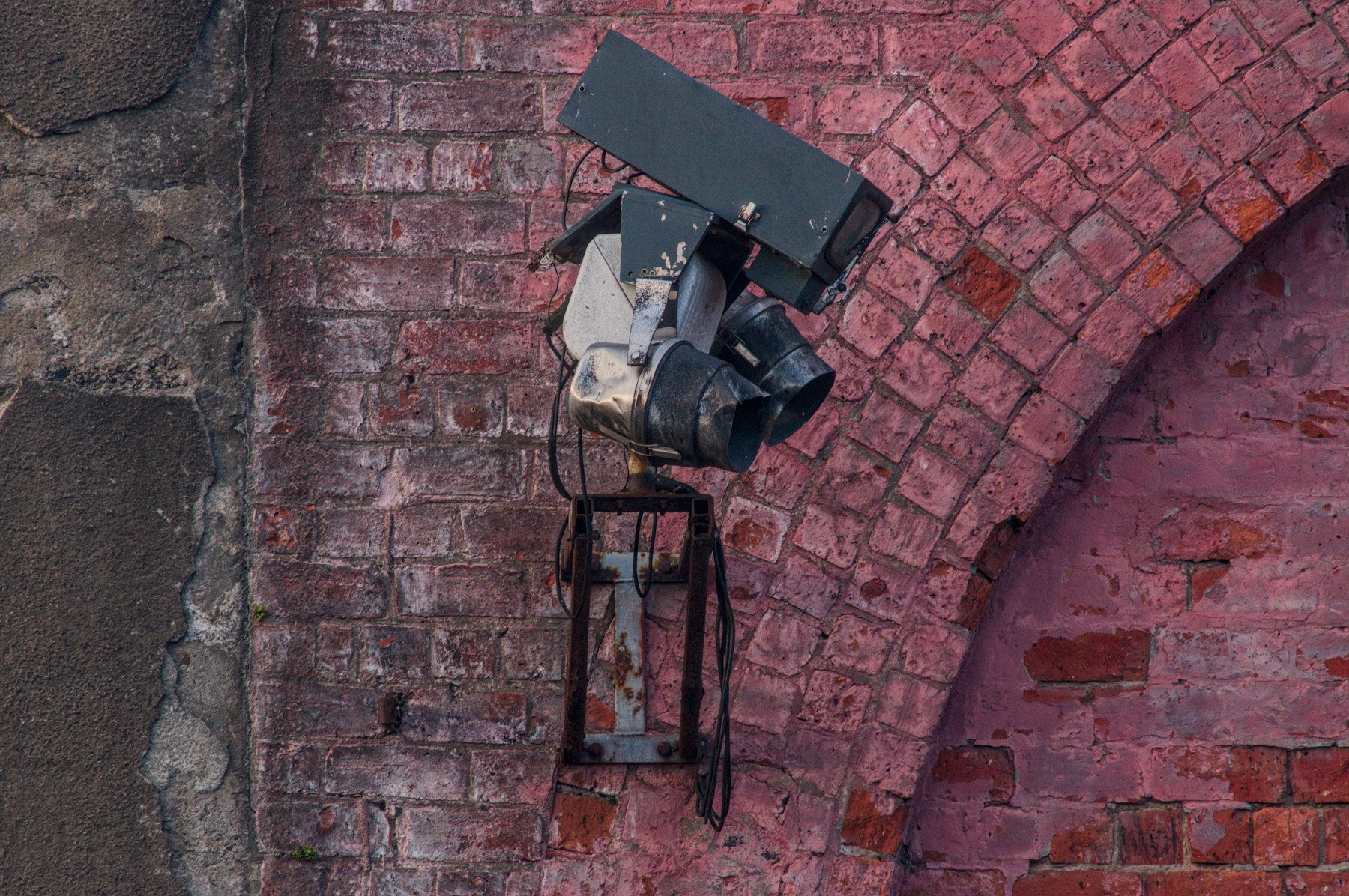 Weathered industrial light fixture with loose cables mounted on a faded red brick wall beside a bricked-up arch at Sentinel Works.