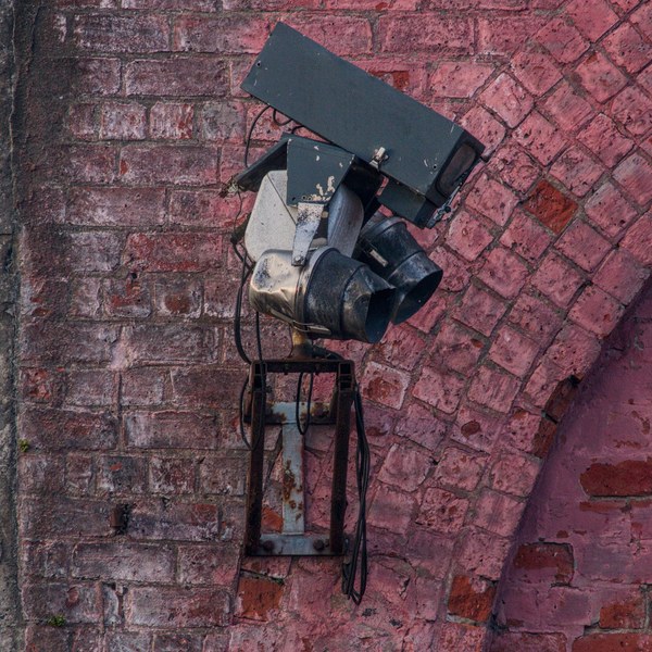 Weathered industrial light fixture with loose cables mounted on a faded red brick wall beside a bricked-up arch at Sentinel Works.
