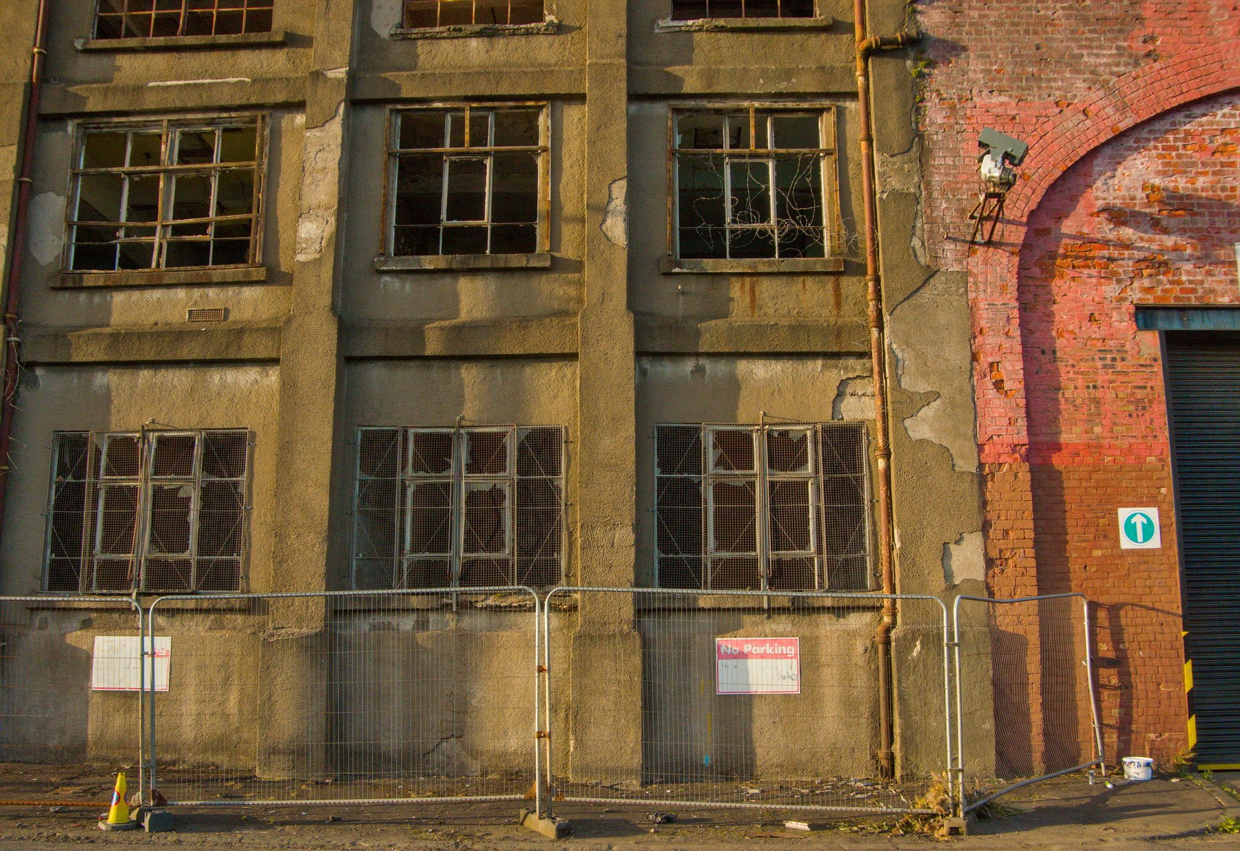 Weathered industrial building façade at Sentinel Works, with broken-paned windows, peeling render, a brick arch, and temporary metal fencing along the pavement.