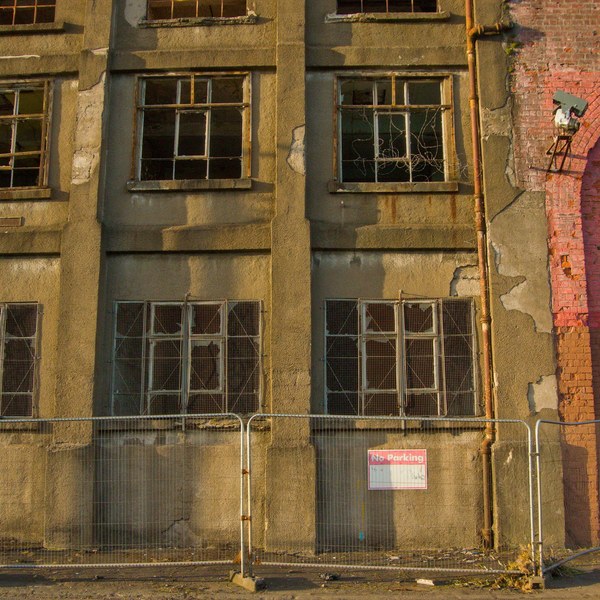 Weathered industrial building façade at Sentinel Works, with broken-paned windows, peeling render, a brick arch, and temporary metal fencing along the pavement.