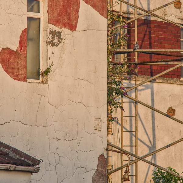Cracked white and red peeling render on an industrial building beside rusted metal scaffolding, with weeds growing through the structure.
