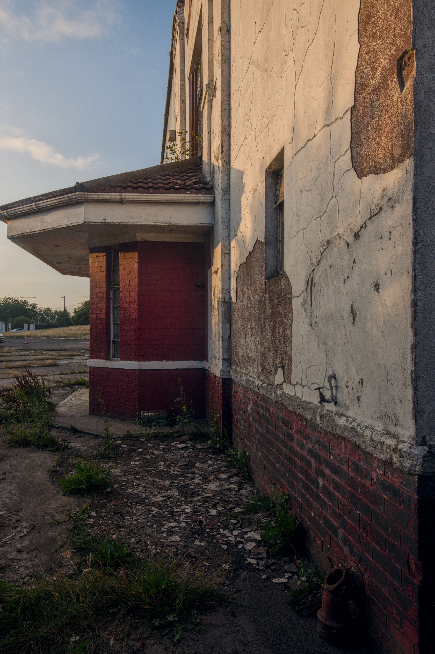 Close view of a derelict industrial building at Shawfield Stadium, with cracked white render, red-brick lower walls and weeds growing beside a boarded entrance.