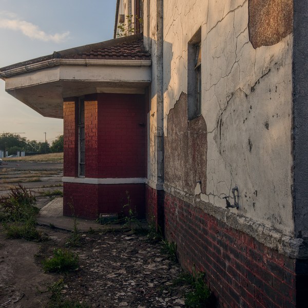 Close view of a derelict industrial building at Shawfield Stadium, with cracked white render, red-brick lower walls and weeds growing beside a boarded entrance.