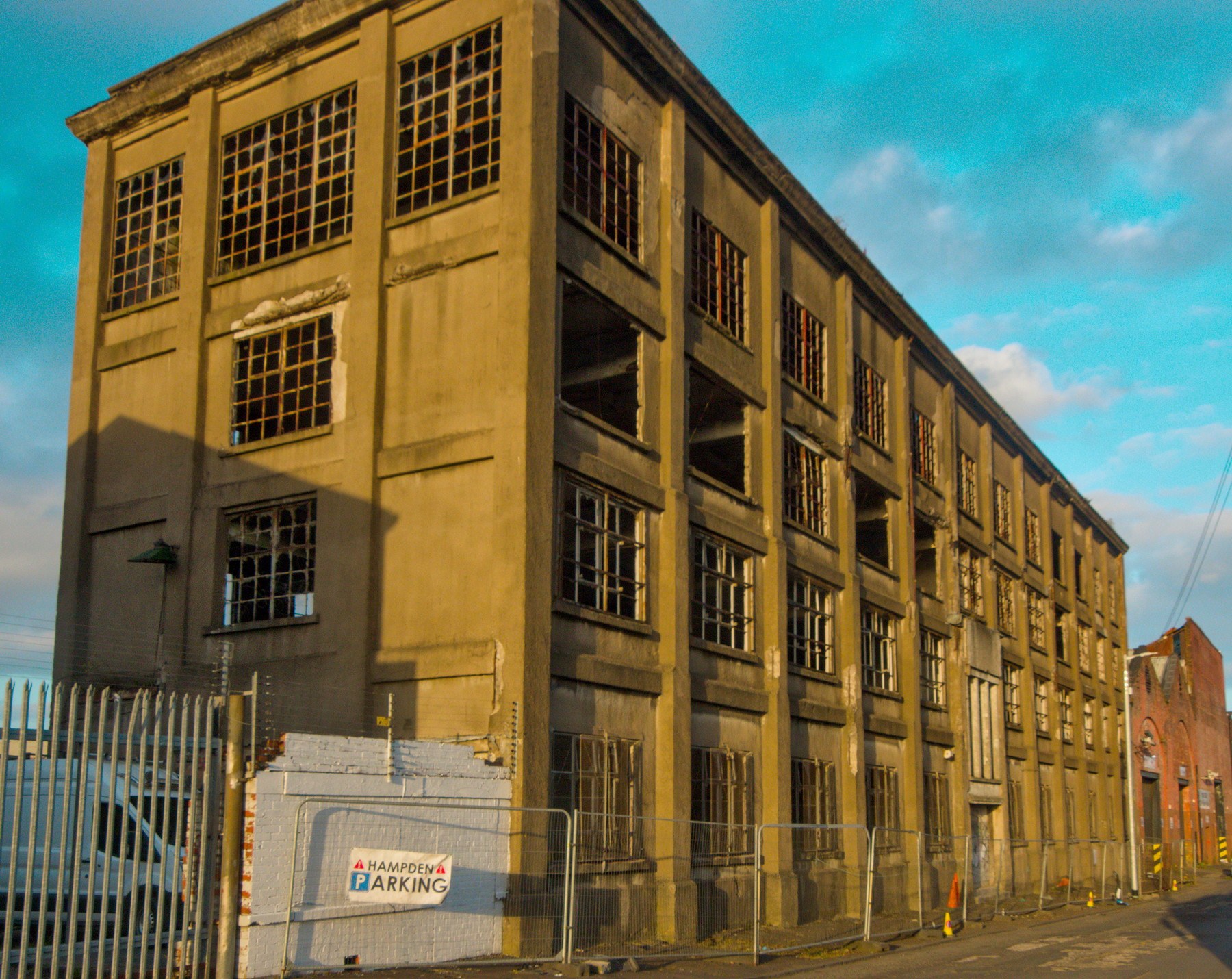 Long abandoned industrial building at Sentinel Works, with rows of broken and empty grid windows above a fenced pavement under a blue, cloud-streaked sky.