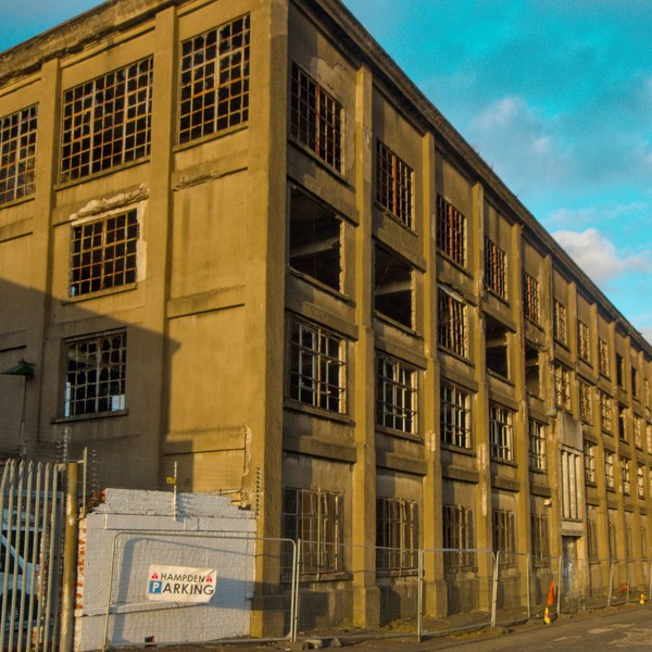 Long abandoned industrial building at Sentinel Works, with rows of broken and empty grid windows above a fenced pavement under a blue, cloud-streaked sky.