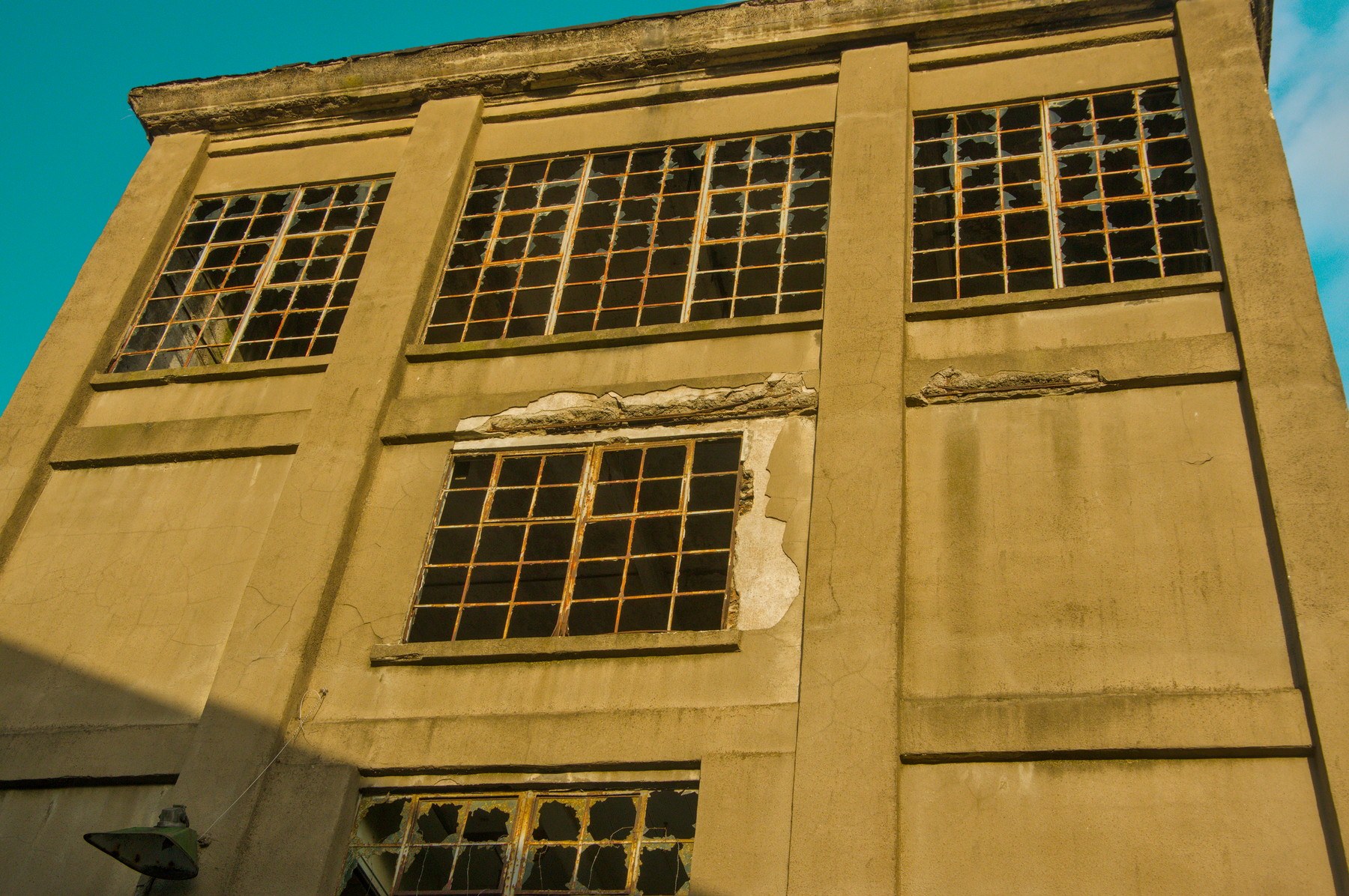 Low-angle view of the weathered concrete facade of Sentinel Works, with large grid windows, many broken panes and cracked, peeling render in warm sunlight against a blue sky.