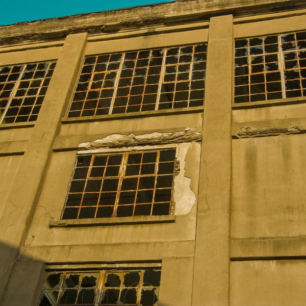 Low-angle view of the weathered concrete facade of Sentinel Works, with large grid windows, many broken panes and cracked, peeling render in warm sunlight against a blue sky.