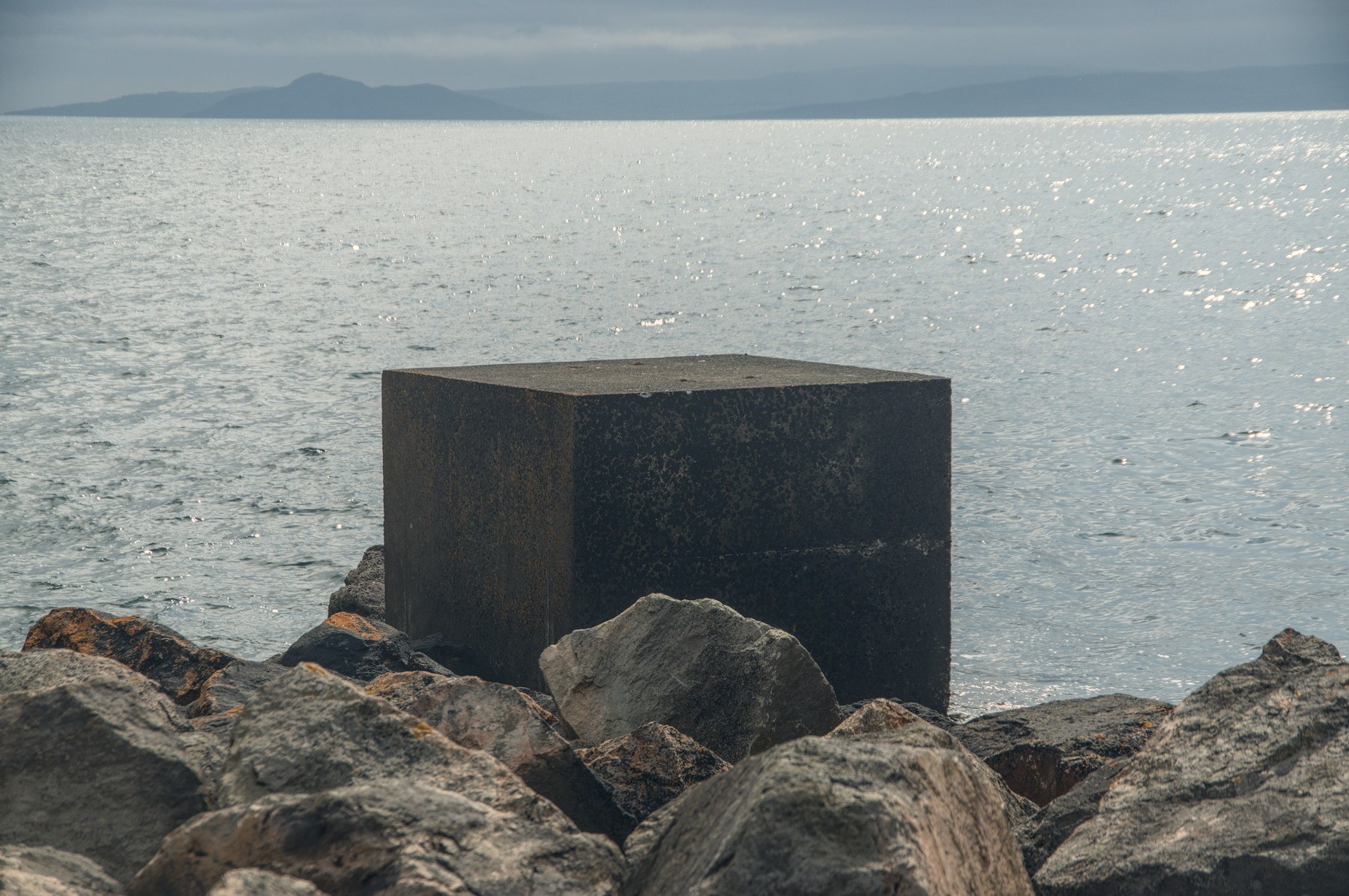 Dark concrete cube standing among large shoreline rocks, with sunlit sea and a hazy island silhouette beyond.