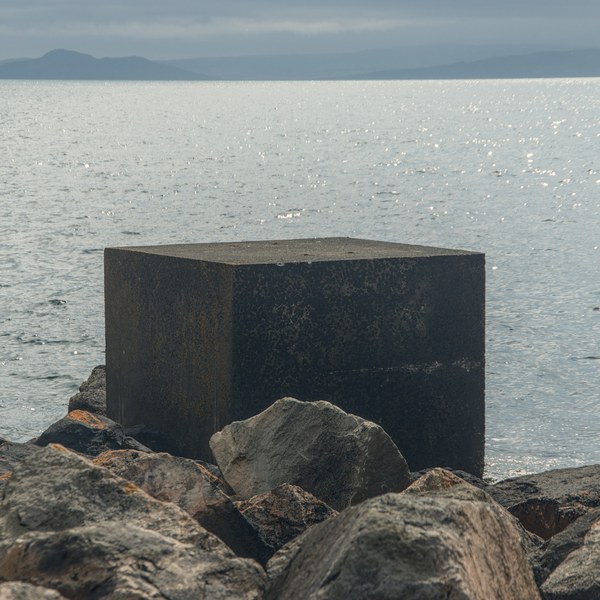 Dark concrete cube standing among large shoreline rocks, with sunlit sea and a hazy island silhouette beyond.