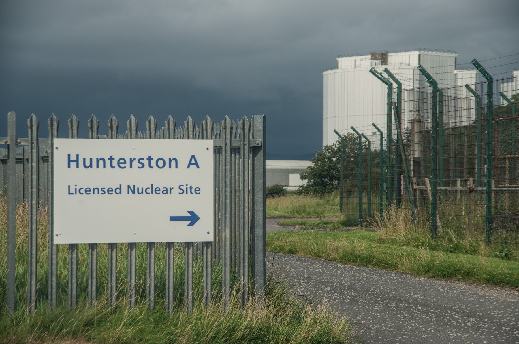 Sign reading “Hunterston A Licensed Nuclear Site” with a right-pointing arrow beside security fencing and large white industrial buildings under dark clouds.