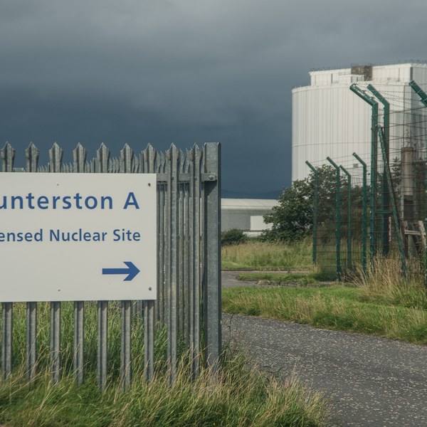Sign reading “Hunterston A Licensed Nuclear Site” with a right-pointing arrow beside security fencing and large white industrial buildings under dark clouds.