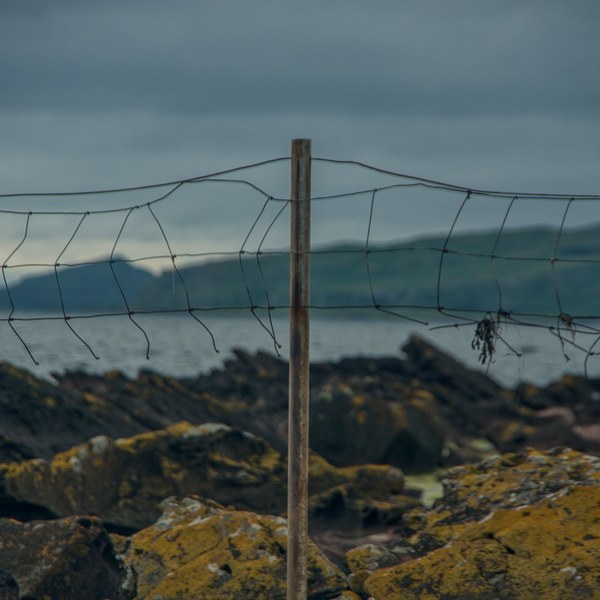 Weathered wire-mesh fence on a wooden post above lichen-covered coastal rocks, with the sea and a dark distant shoreline beneath heavy clouds at Portencross, Ayrshire.