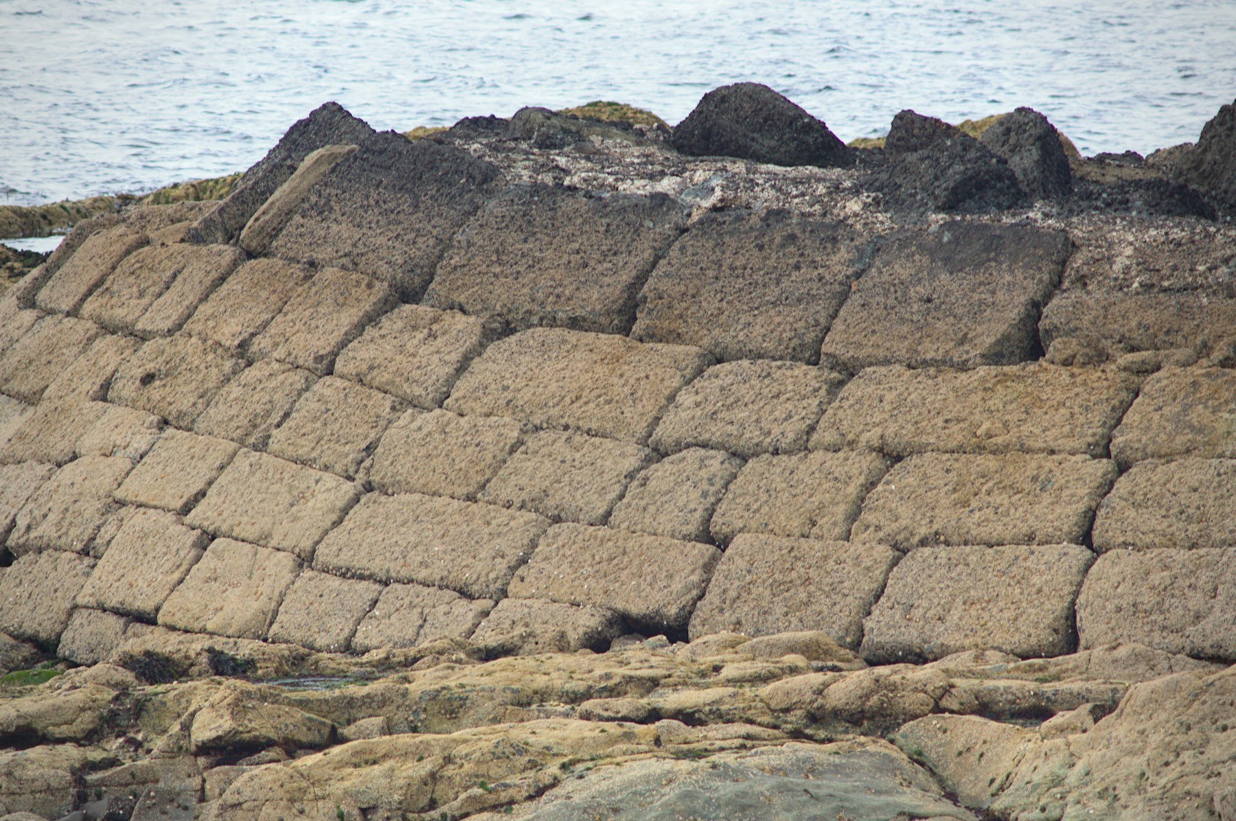 Weathered sloping stone retaining wall made of large rectangular blocks, with dark rocks along its top and the sea behind.
