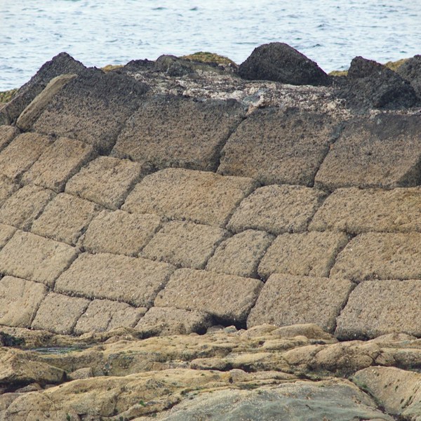 Weathered sloping stone retaining wall made of large rectangular blocks, with dark rocks along its top and the sea behind.