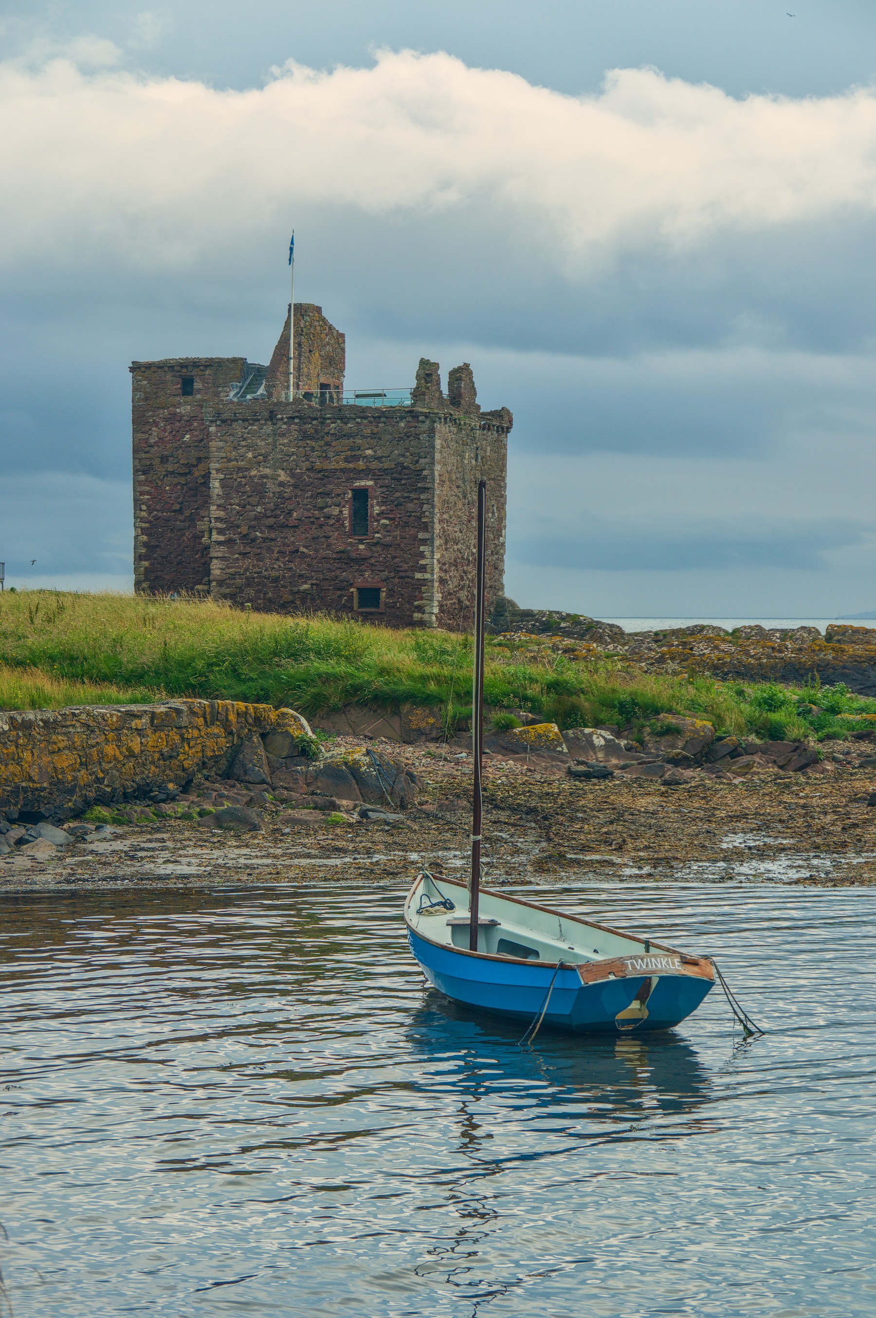Blue sailing dinghy moored on calm water below a roofless stone coastal ruin at Portencross, beneath a cloudy sky.