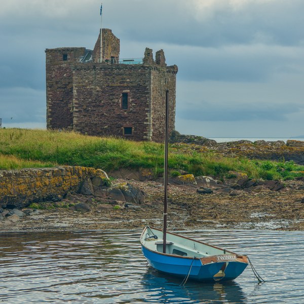Blue sailing dinghy moored on calm water below a roofless stone coastal ruin at Portencross, beneath a cloudy sky.